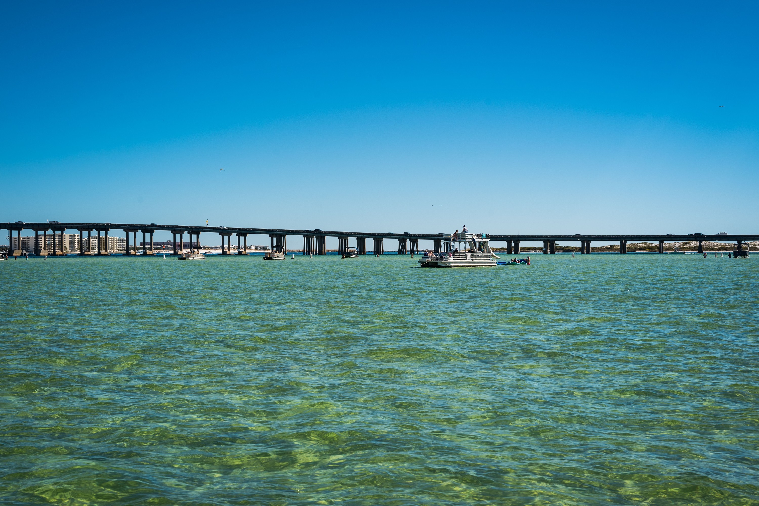 A double-decker pontoon boat on emerald water in front of the Destin bridge, seen from the water at Crab Island