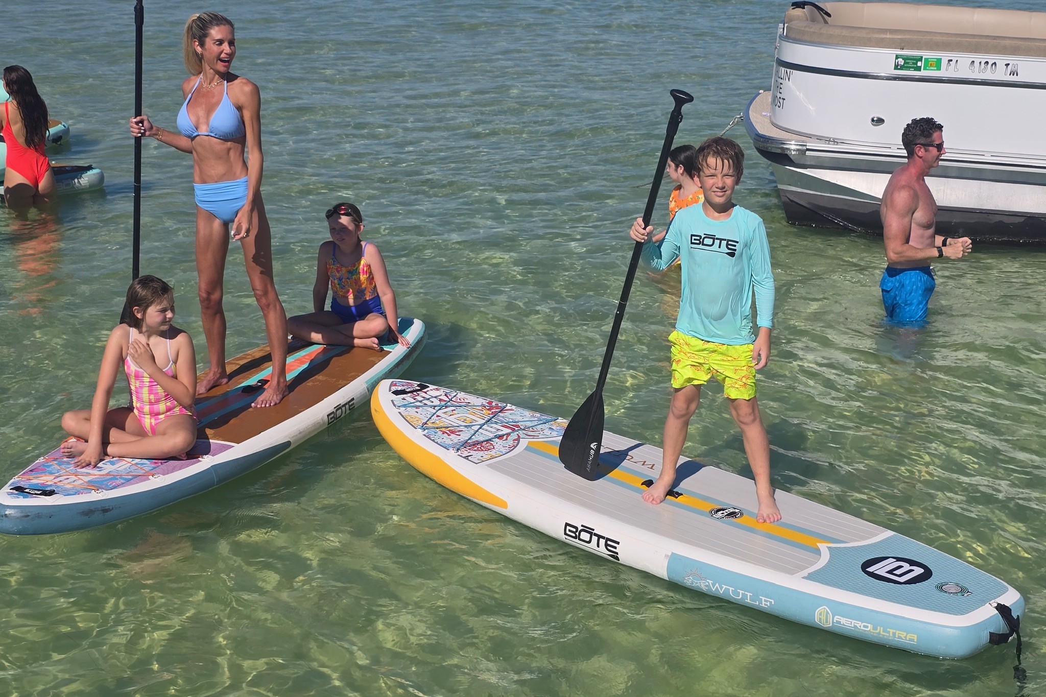 A family paddleboarding in shallow clear water at Crab Island, boats anchored behind them