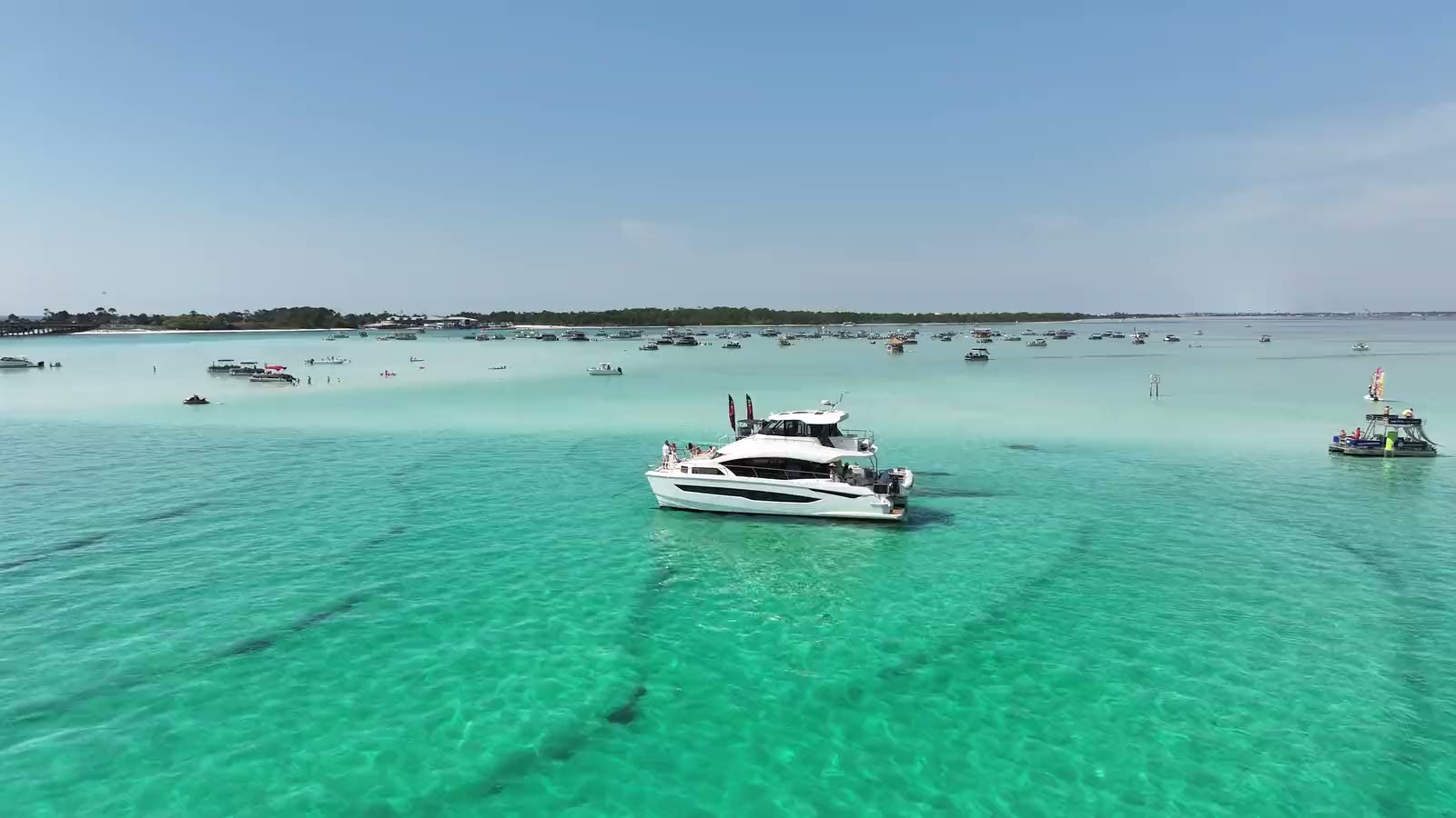 A yacht at the Crab Island sandbar with boats anchored across it, the Destin bridge and skyline behind, seen from above
