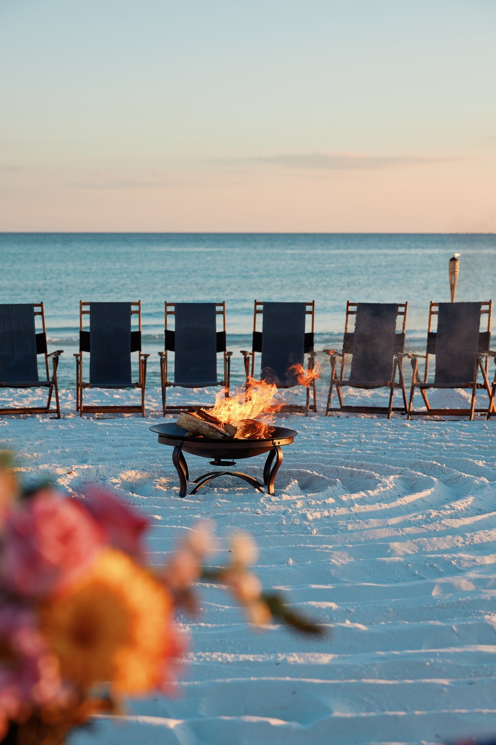 A bonfire on the sand at sunset, with lounge chairs and flowers