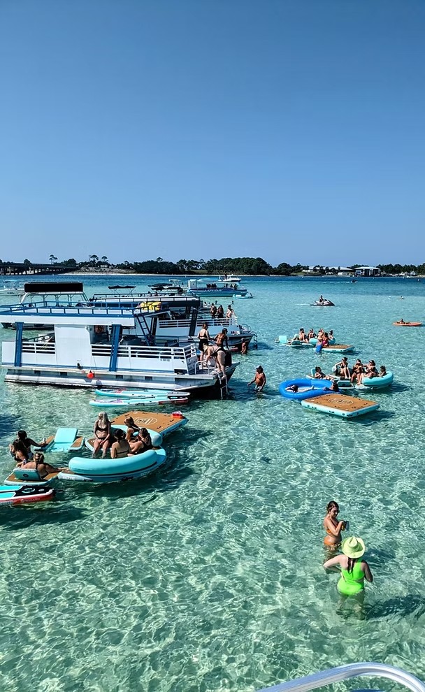 People wading and floating in clear water at the Crab Island sandbar, boats anchored behind them