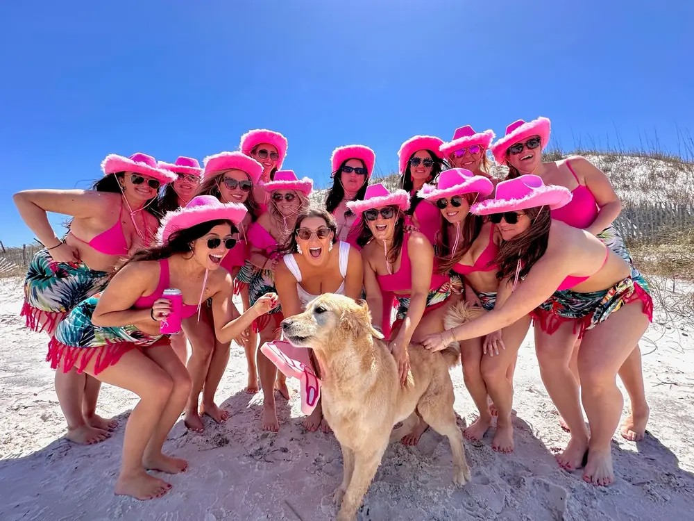 A group in pink cowboy hats with a golden retriever on a white-sand beach