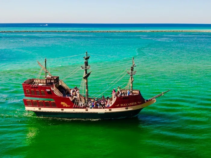 A red-hulled pirate ship underway on emerald water, seen from above