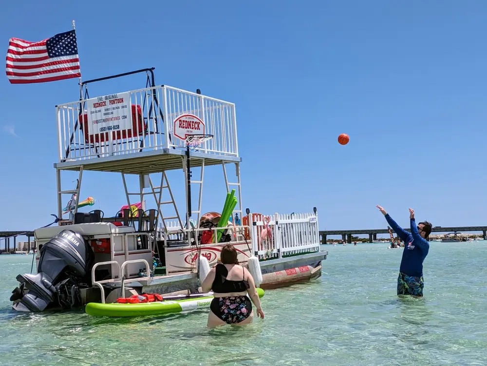 A double-decker pontoon flying a flag, with a basketball hoop and people playing in the shallows